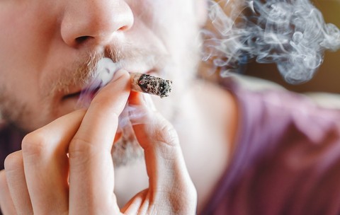 Close-up cropped shot of young man smoking marijuana or cannabis joint indoors, male hands with cigarette, home environment, lifestyle, addiction and recreational use concept. 