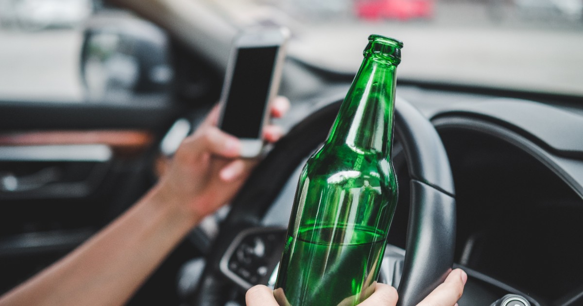 Un bărbat care conduce o mașină, dar într-o mână ține un telefon mobil și în altă mână o sticlă de bere