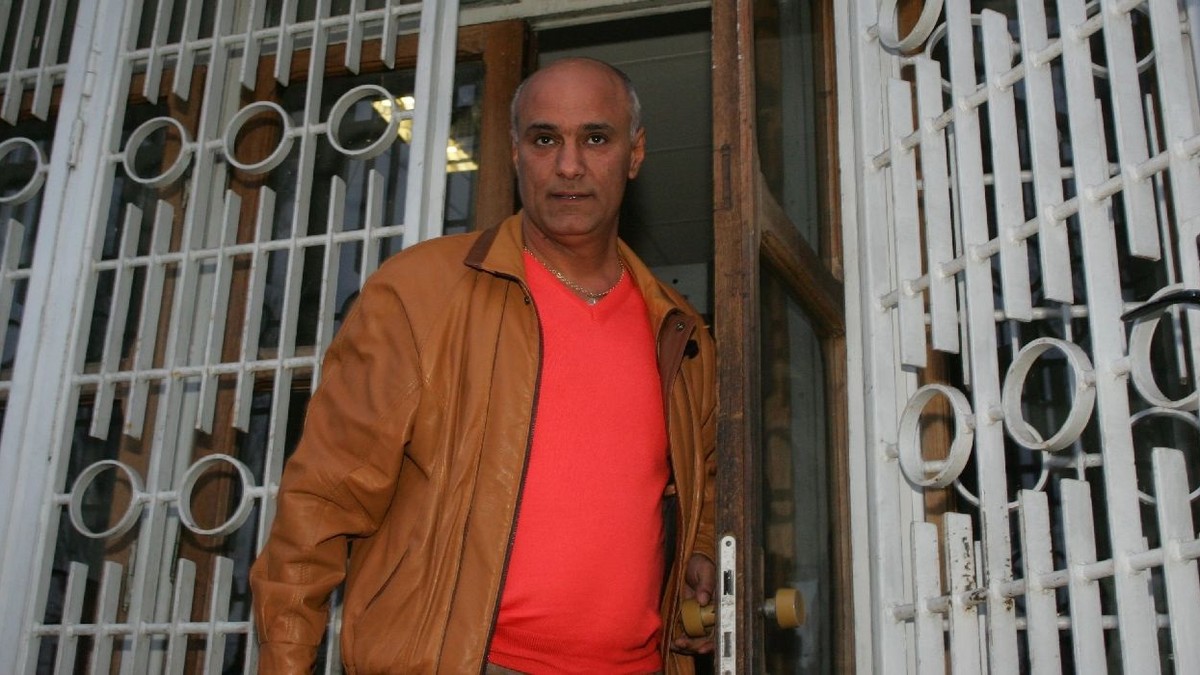 Un bărbat cu geacă maro și tricou portocaliu iese pe o poartă din fier