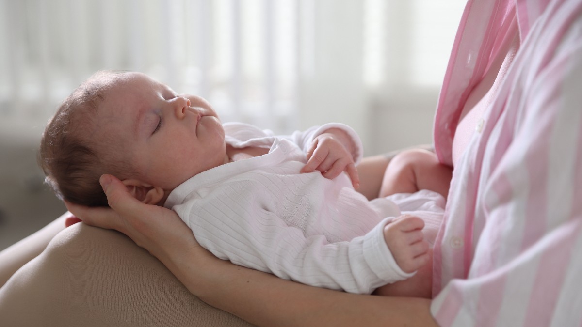 Imaginea prezintă un bebeluș care doarme liniștit, culcat pe brațele și pe picioarele unei persoane (văzute în planul apropiat). Bebelusul, îmbrăcat în alb, este ținut cu grijă în lumină naturală, creând o scenă de tandrețe și confort.