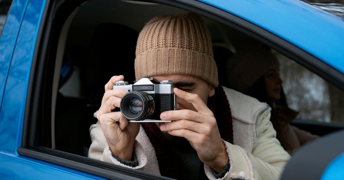 bărbat cu căciulă și haine crem, într-o mașină albastră cu geamul deschis, ținând în fața feței un aparat de fotografiat