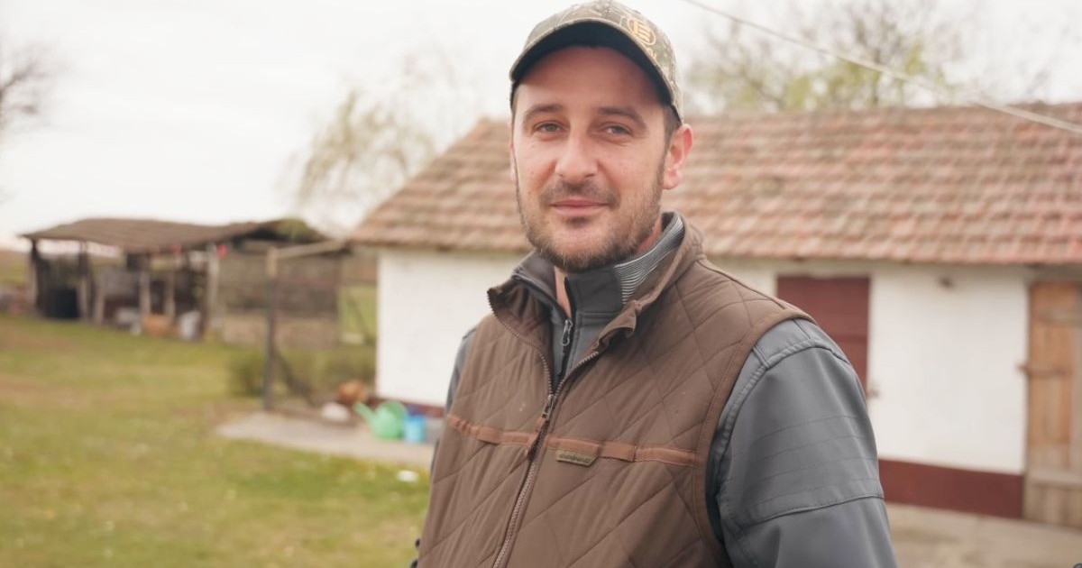 Imaginea surprinde un tânăr care privește direct spre cameră și zâmbește ușor. Poartă o șapcă de camuflaj, o vestă maro peste o jachetă gri și se află într-o zonă rurală. În fundal se vede o casă tradițională cu pereți albi și acoperiș din țiglă.