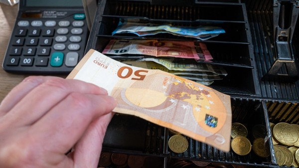 O mână care pune o bancnotă de 50 de euro într-un sertar deschis al unei case de marcat, lângă compartimentele pline cu monede și bancnote euro.