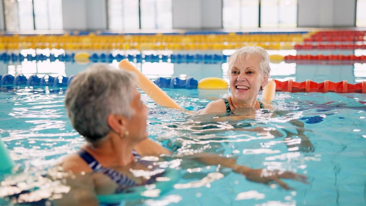 Două persoane în vârstă în timp ce înoată și fac sport medical în piscina dela băi, sunt vesele și zâmbesc