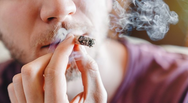Close-up cropped shot of young man smoking marijuana or cannabis joint indoors, male hands with cigarette, home environment, lifestyle, addiction and recreational use concept. 