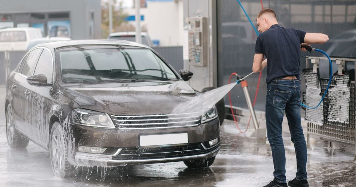 Un bărbat îmbrăcat cu blugi negri și o bluză neagră cu mâneci lungi spală o mașină de culoare maro închis, la o spălătorie auto