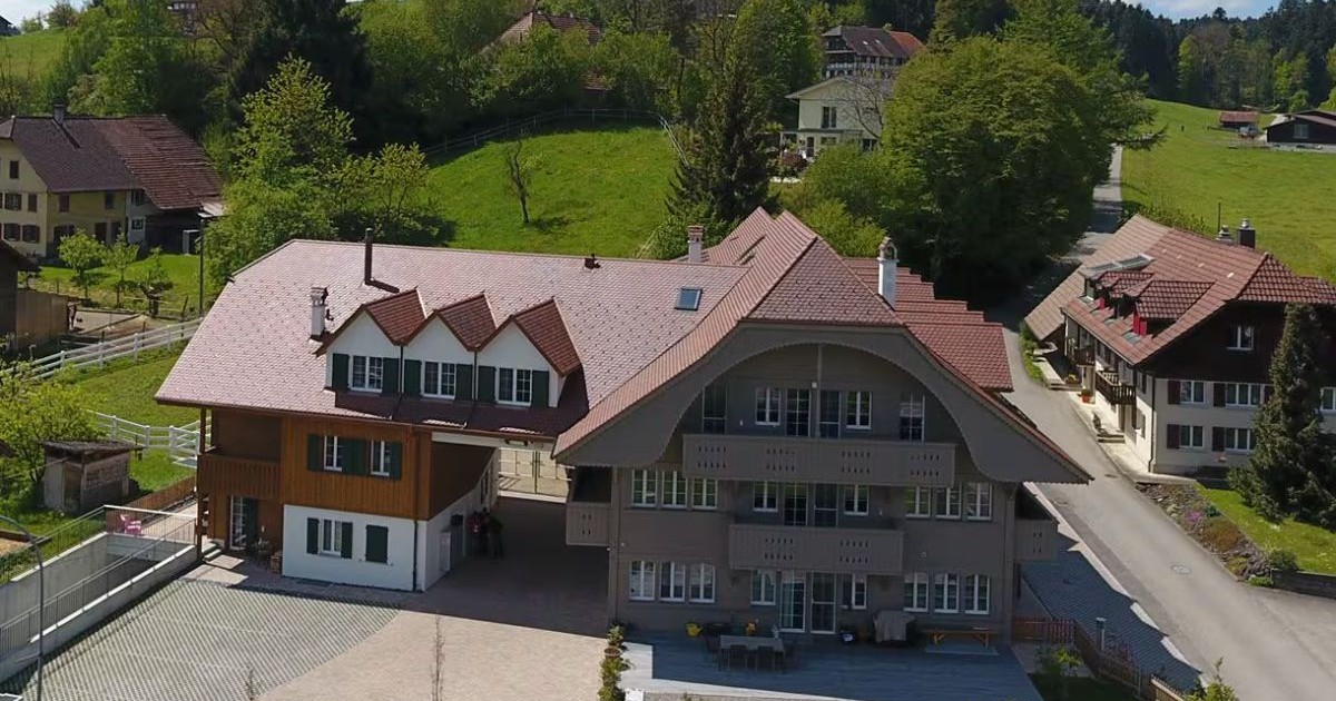 Clădirea fermei, cu acoperiș de culoare maro închis, înconjurată de pădure și pășune