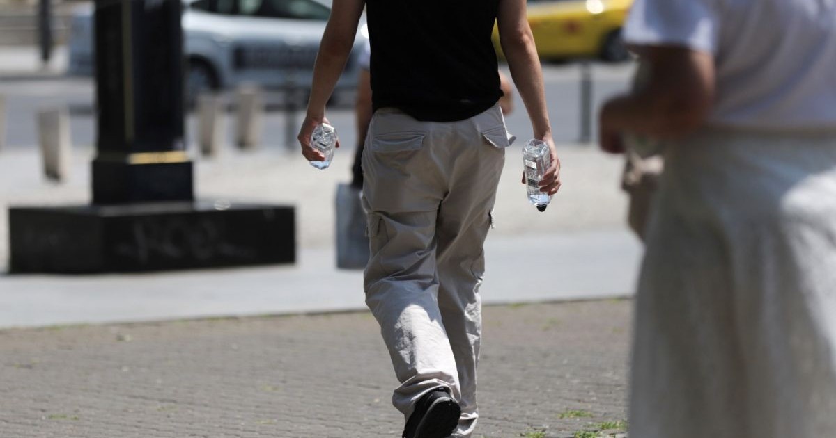 Un tânăr îmbrăcat cu tricou negru și pantaloni deschiși la culoare, fotografiat cu spatele, ține în fiecare mână câte o sticlă de apă, într-o zi caldă de vară, București, august 2025