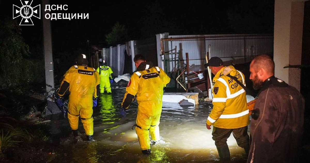 Imaginea surprinde un grup de salvatori îmbrăcați în echipamente de protecție galbene reflectorizante, intervenind pe timpul nopții într-o zonă inundată. Bărbații pășesc prin apă tulbure care le ajunge până la genunchi, sugerând o operațiune de salvare în urma unei furtuni sau inundații în Odesa.