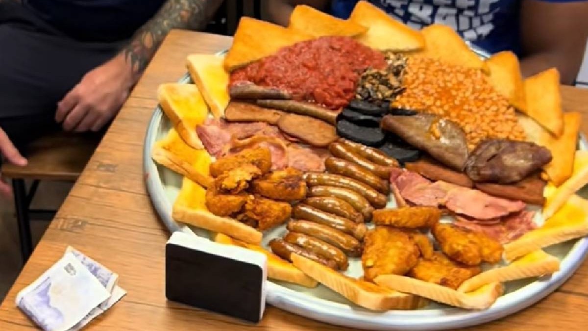 Un patou urias cu mancare de tip fast food, printre care 10 cârnați, mai multe felii de bacon, ciuperci, roșii, fasole, budincă neagră, opt cartofi prăjiți și 16 felii de pâine prăjită, asezat pe o mas[