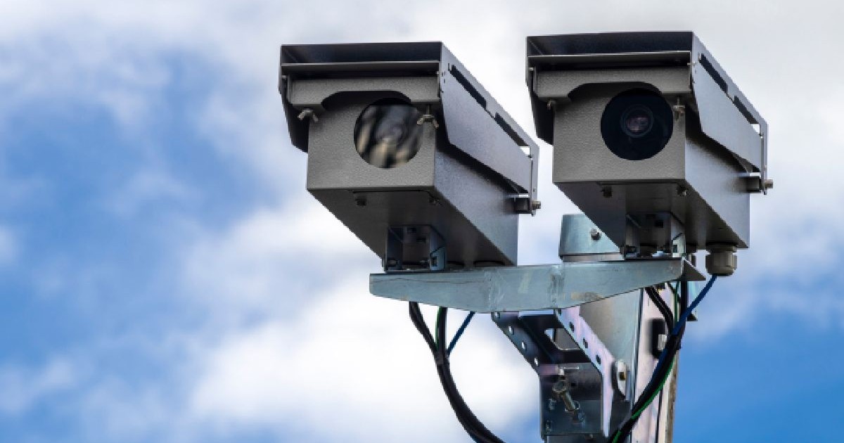 Fotografie cu două camere radar de control al vitezei, montate pe un stâlp metalic, pe fundalul unui cer albastru parțial înnorat.