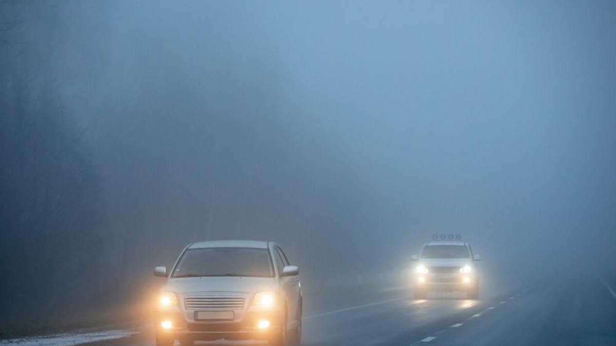 Două mașini cu farurile aprinse, una în spatele alteia, merg pe autostradă într-o dimineață cu ceață