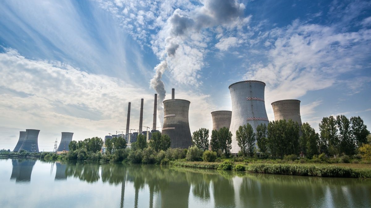 Termocentrala Turceni, cu un lac în față, turnurile uriașe pe fundal pe care iese fum și cerul albastru cu nori albi