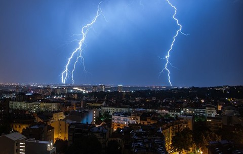 Furtună cu fulgere și cer înnorat peste un oraș, cu două fulgere care lovesc pământul în fundal