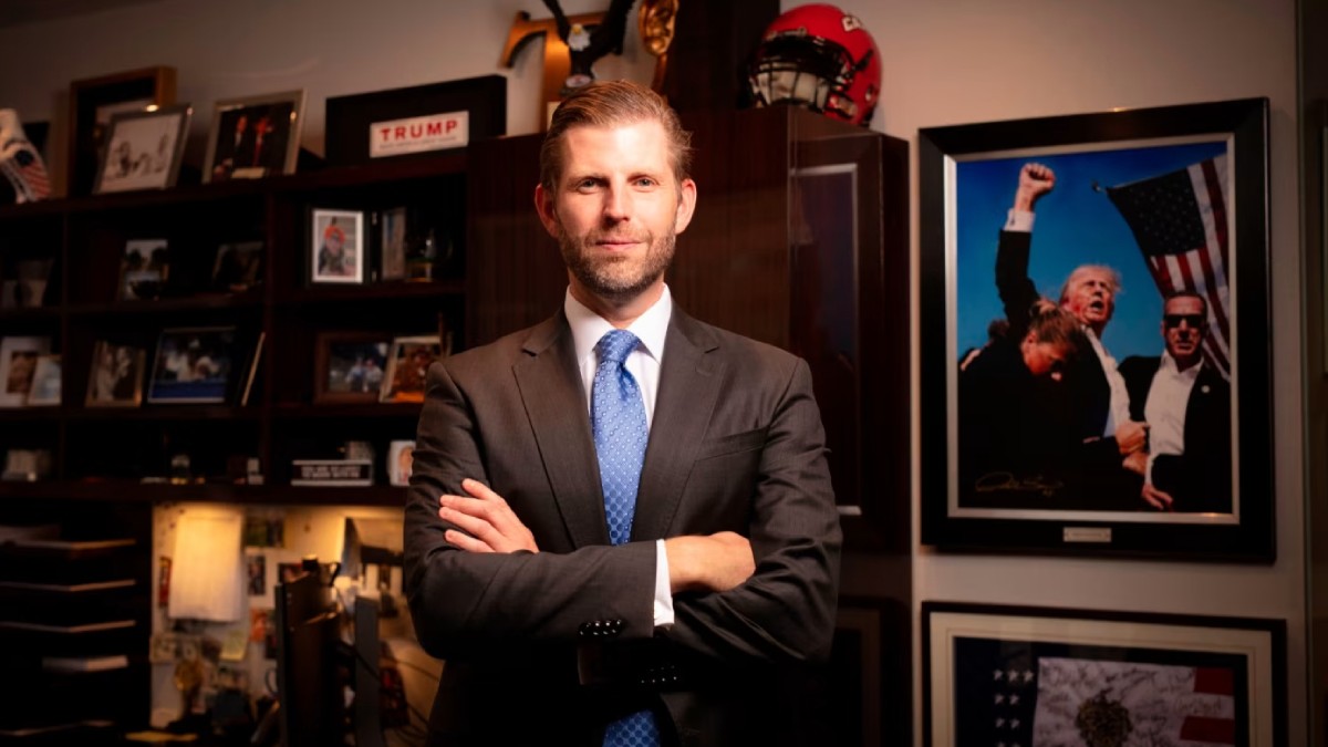 Eric Trump, fiul lui Donald Trump, îmbrăcat într-un costum negru, cămașă albă și cravată bleu, cu mâinile încrucișate la piept. În spatele său, în dreapta, o fotografie înrămată pe perete cu Donald Trump după prima tentativă de asasinat. În spatele său, în stânga, o bibliotecă în care sunt înrămate mai multe fotografii.