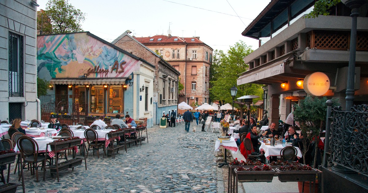 O stradă pietruită, plină de farmec, este mărginită de clădiri vechi și restaurante cochete, unde oamenii stau la mese în aer liber și se bucură de atmosferă. Luminile calde și decorul rustic creează un aer boem, amintind de vremuri mai liniștite și de tradițiile locului.