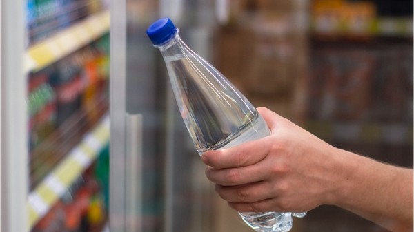 O mână care ține o sticlă din plastic cu apă și un capac albastru, scoasă sau așezată lângă rafturile frigorifice ale unui magazin.