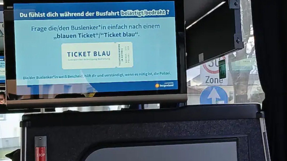 Imaginea arată un ecran montat în interiorul unui autobuz, pe care este afișat un mesaj informativ despre „Ticket Blau” (biletul albastru). Anunțul explică faptul că pasagerii care se simt hărțuiți sau amenințați pot cere șoferului un „bilet albastru” pentru a primi ajutor în mod discret.