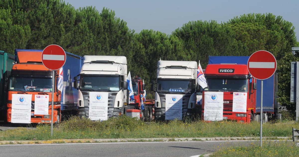Mai multe camioane de transport staționează într-o parcare din Italia, în cadrul unei acțiuni de protest organizate de transportatori. Vehiculele sunt aliniate cu bannere și steaguri afișate în față, într-o atmosferă de mobilizare și solidaritate profesională.