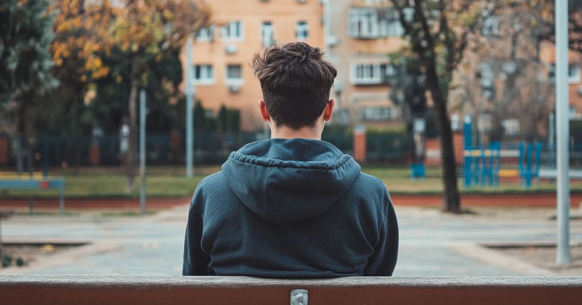 adolescent care stă pe o bancă de lemn maro, îmbrăcat într-un hanorac verde închis, cu spatele. în fundal se văd clădiri galbene și portocalii și copaci verzi