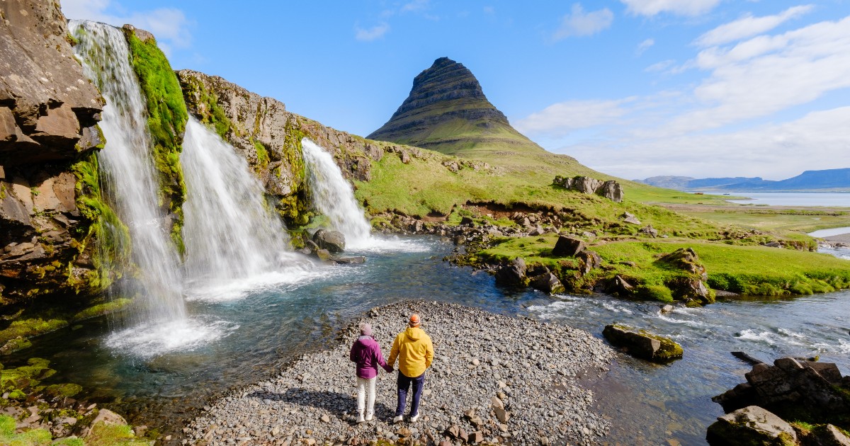 Imaginea surprinde un peisaj vulcanic spectaculos din Islanda, dominat de cascada Kirkjufellsfoss (Cascada Muntelui Biserică) și de conul izolat al muntelui Kirkjufell pe fundal. Un cuplu, îmbrăcat în jachete de drumeție, stă cu spatele la cameră pe o zonă pietroasă, admirând priveliștea.