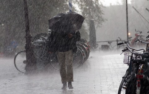 O persoană care merge pe un trotuar în timpul unei ploi torențiale cu vânt, ținând o umbrelă neagră aplecată în față, pe lângă un rând de biciclete parcate.
