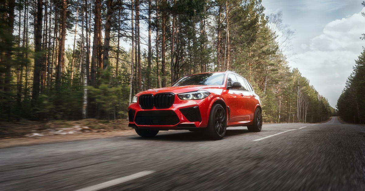 Mașina BMW X5 de culoare roșie se deplasează pe autostradă într-o zonă cu pădure.