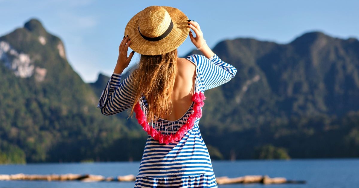 Young pretty blonde woman with long hairs, wearing vintage hat and bright sexy trendy dress looks to the mountains and lake, show her hand, amazing summer adventures.