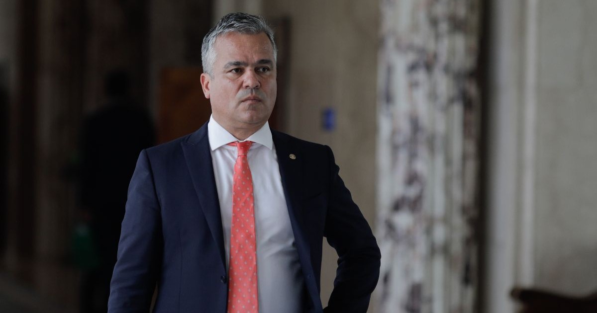 Adrian Veștea, îmbrăcat în costum închis la culoare, cămașă albă și cravată portocalie, mergând pe un coridor, cu mâna dreapta în buzunar