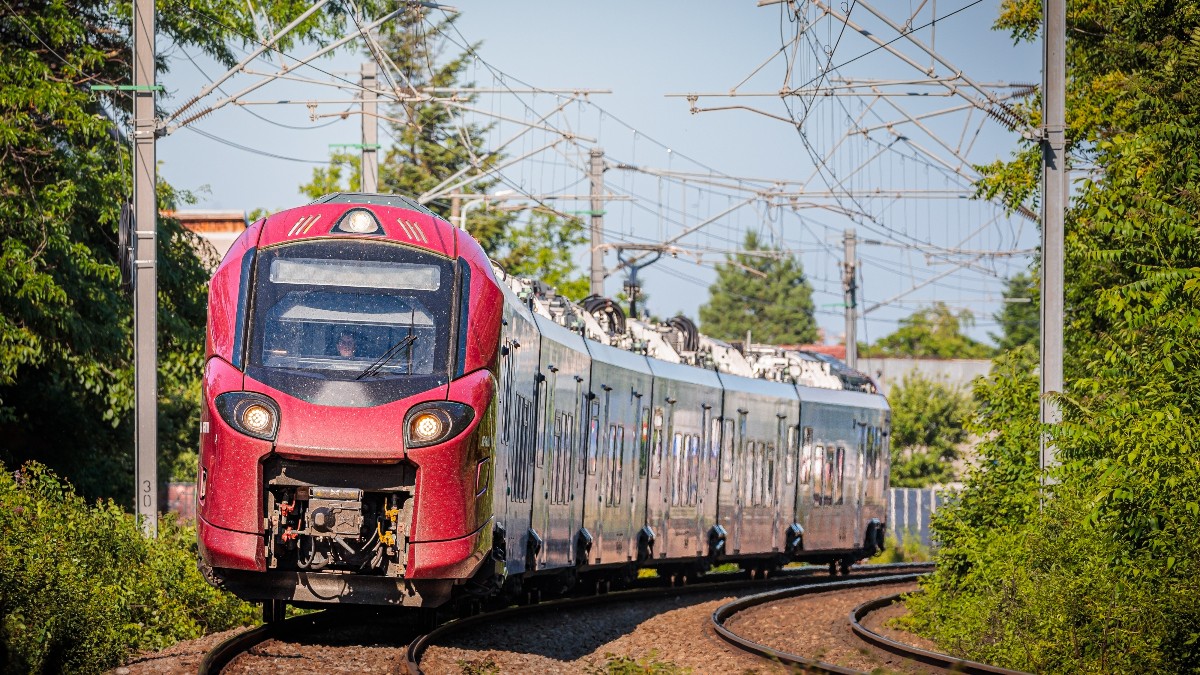 Imaginea prezintă un tren modern, de culoare roșie și argintie, care circulă pe o linie ferată curbată. În jur se observă vegetație verde și stâlpii rețelei electrice, sugerând un traseu feroviar în aer liber, într-o zonă liniștită.