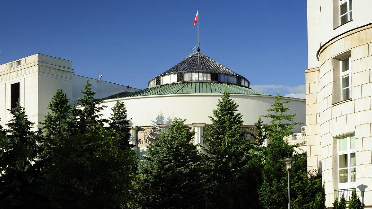 În imagine e Parlamentul Poloniei, înconjurat de brazi înalți și cu un steag pus în vârful clădirii