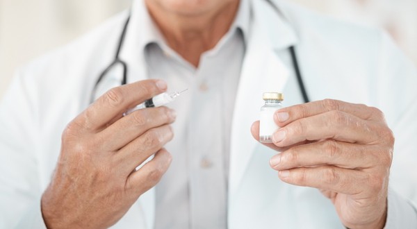 Un doctor îmbrăcat în halat alb, purtând un stetoscop în jurul gâtului, care ține o seringă cu ac și un flacon mic de sticlă