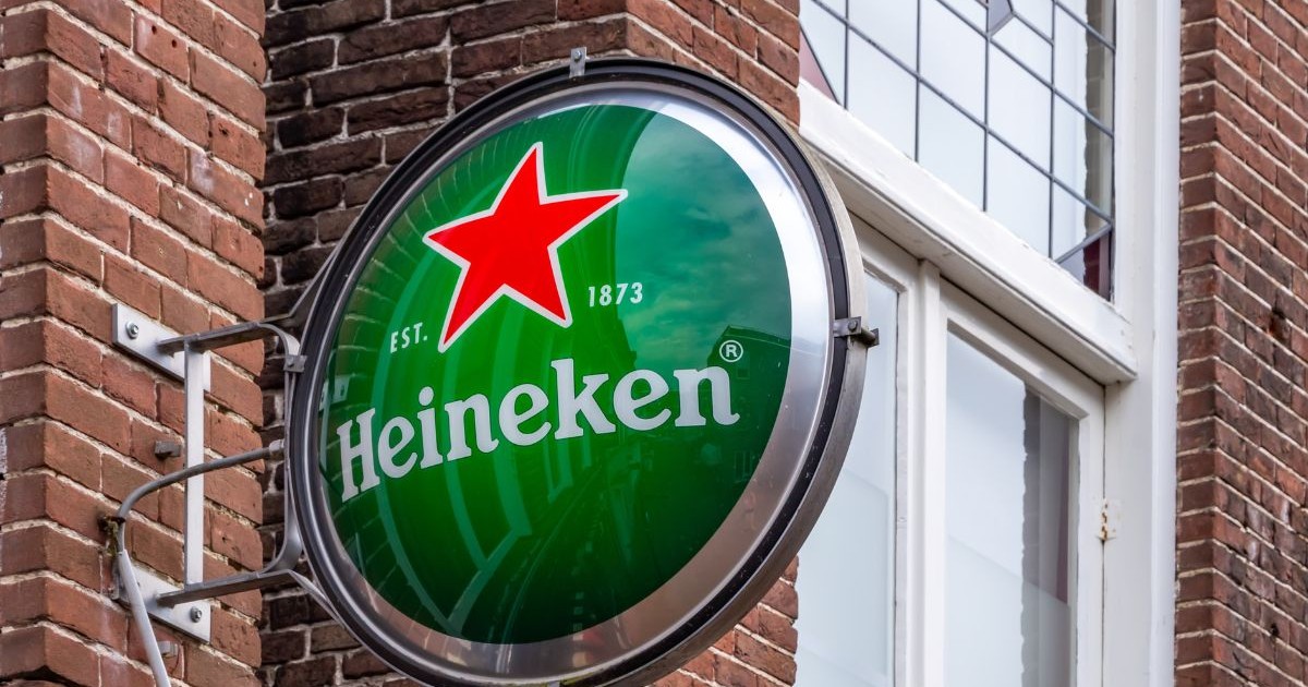 Sigla lucioasă și rotundă a brandului „Heineken”, montată perpendicular, pe un perete de cărămidă tradițional.