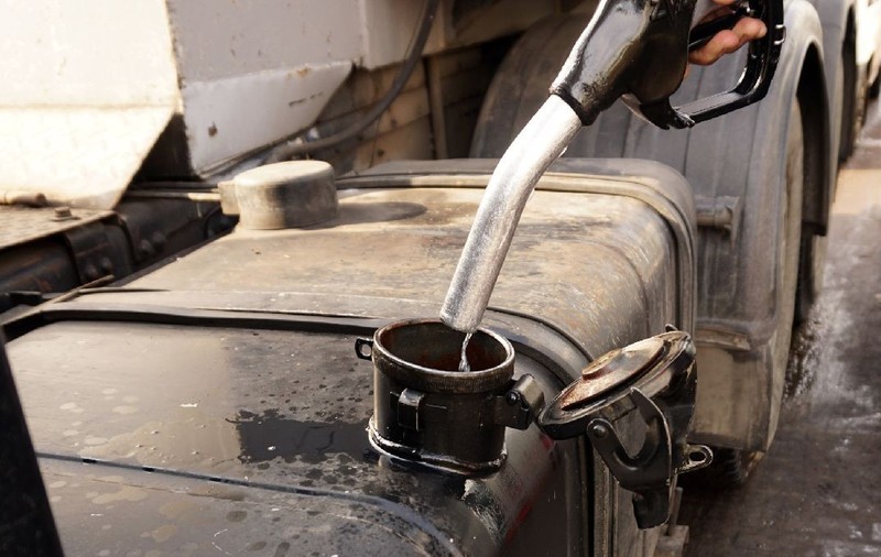 Prim-plan al unei persoane care toarnă motorină dintr-o pompă de combustibil neagră într-un rezervorul unui camion. În fundal se văd șasiul metalic și anvelopele vehiculului greu. Sursa foto: Shutterstock