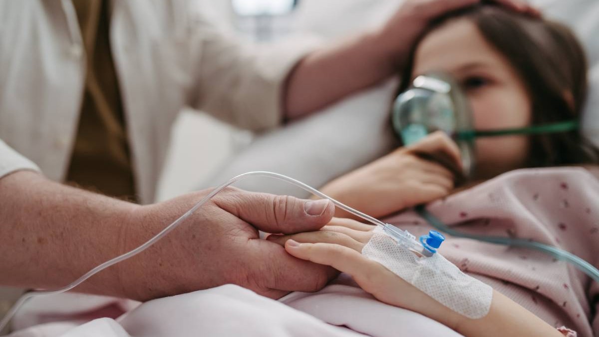 Mana unui barbat in halat alb tine de mana o fetita, aflata pe patul de spital, cu o masca de oxigen pe fata,