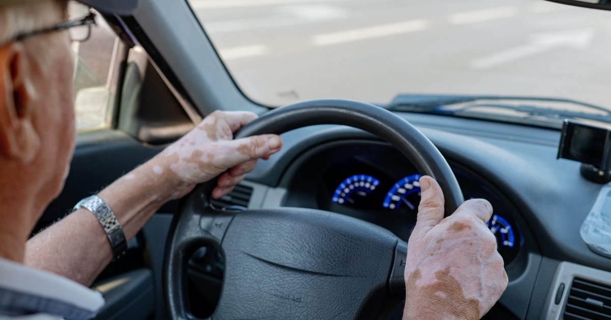 În imagine se vede o persoană în vârstă aflată la volanul unei mașini. Mâinile sale sunt poziționate pe volan, iar în față se observă bordul și drumul pe care circulă.