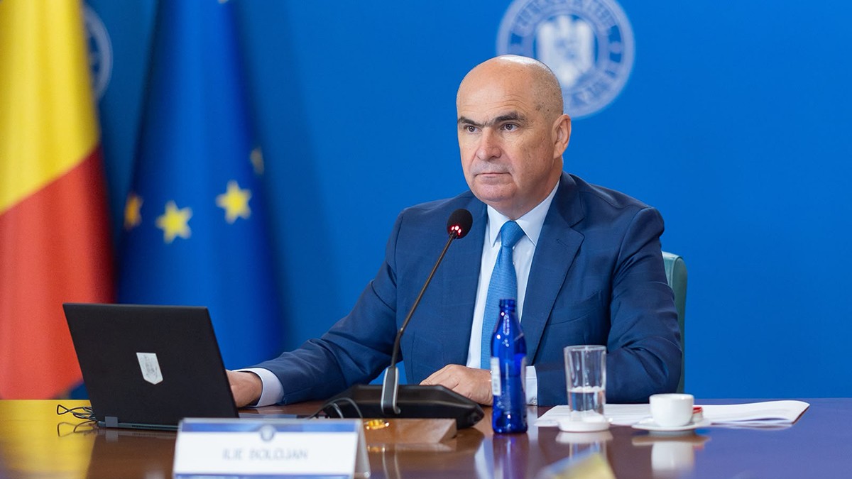 Premierul Ilie Bolojan sta la un birou avand steagul Romaniei si Ue in partea stânga