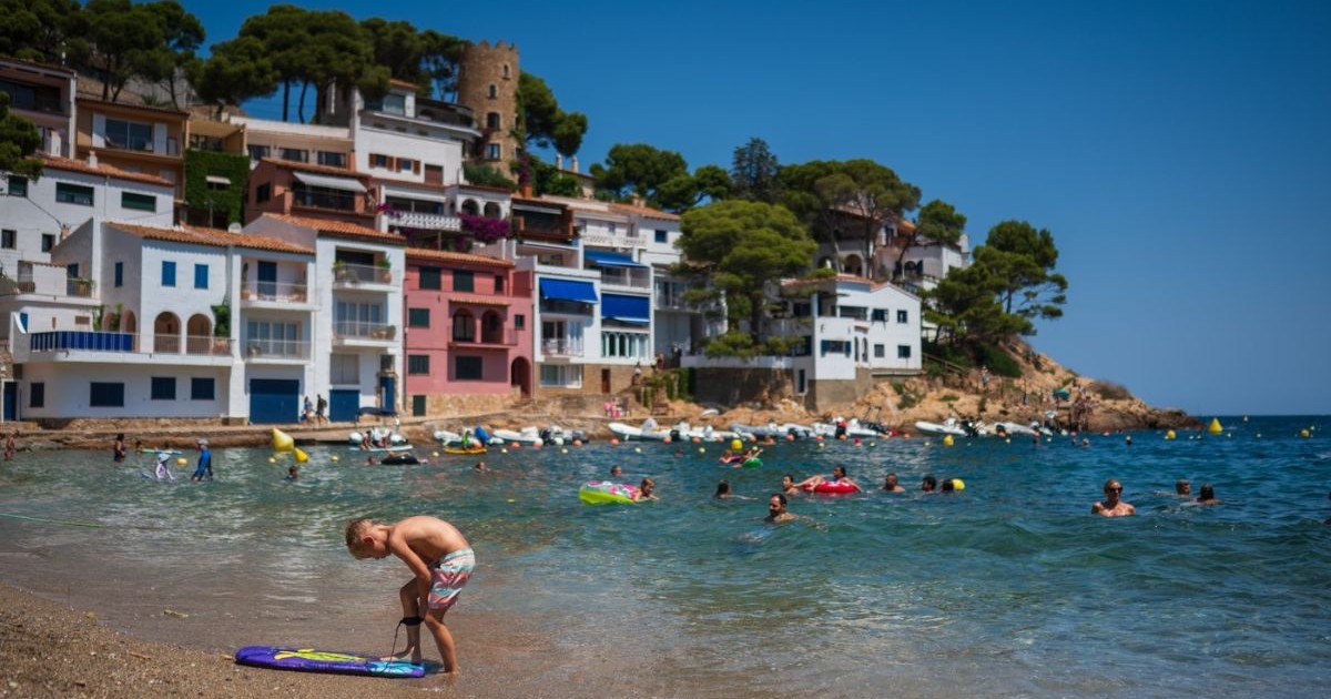 o plajă cu nisip, unde sunt persoane atât în apă, cât și pe mal. În centrul imaginii se află un copil în costum de baie, aplecat lângă o placă de surf mică. Apa este de culoare albastru-verde, cu valuri mici, iar oamenii din apă folosesc materiale gonflabile colorate. În fundal, pe versantul unei coline, se ridică mai multe case cu fațade albe, roz, albastre și balcoane largi, înconjurate de pini și vegetație verde