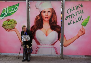 epa03318719 An elderly Russian beggar woman stands in front of billboard showing famous Russian figure skater Anna Semenovich advertising for ice cream in Moscow, Russia, 26 July 2012. The begging woman holds a placard asking passers-by to help her to buy bread. EPA/SERGEI CHIRIKOV