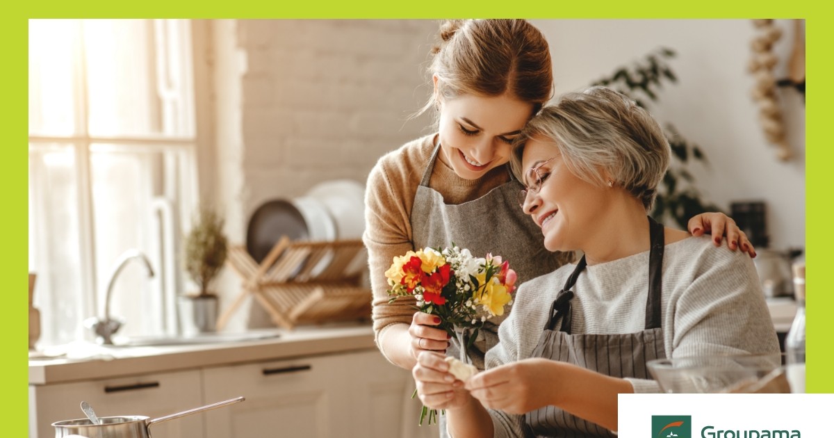 O mama impreuna cu fiica. Mama, cu parul carunt sta jos pe scaun, sprijinindu-si capul de umarul fiicei ei, care ii ofera un buchet de flori