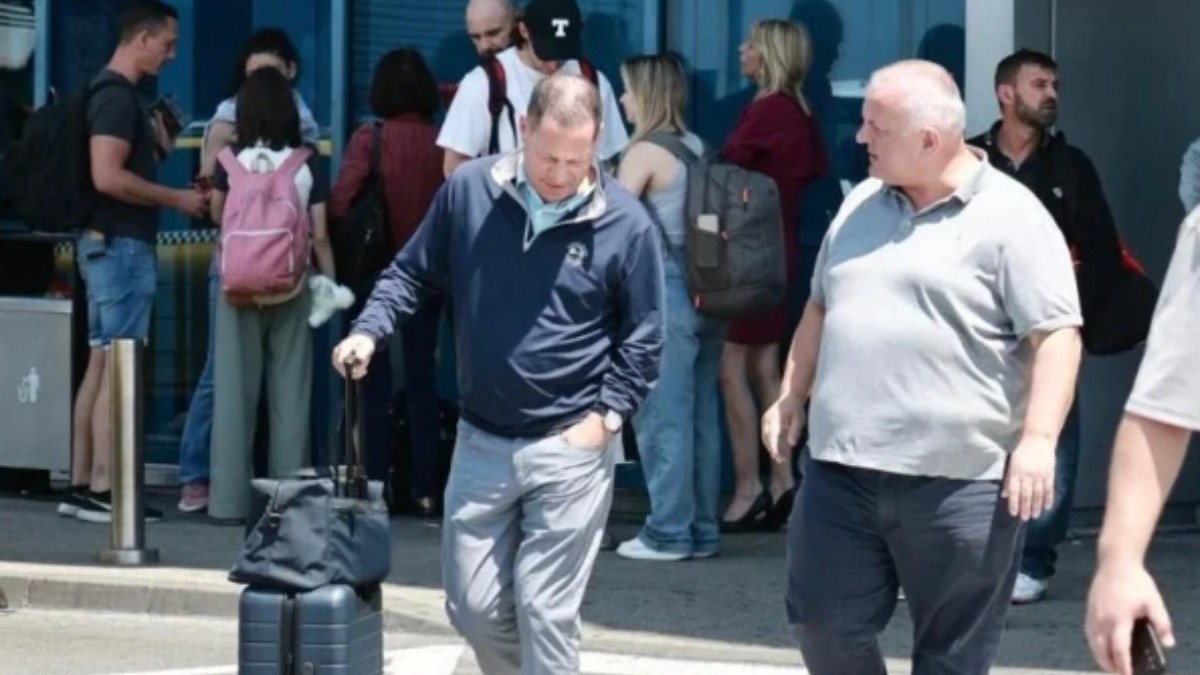 În prim-plan se află doi bărbați: unul cu un troller și o geantă de mână, îmbrăcat într-o jachetă sport și pantaloni deschiși la culoare, care a aterizat recent; celălalt poartă un tricou gri și pantaloni închiși la culoare, și îl privește pe primul.