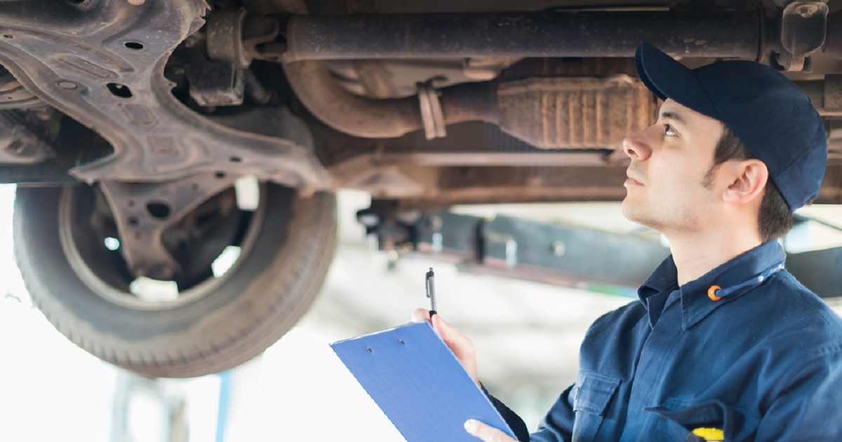Ce este flitrul de particule al unei maşini şi cum se întreţine - Un mecanic auto verifică partea inferioară a unei masini, ridicată pe elevator, într-un service, ținând în mână un dosar clipboard cu documente pentru notițe