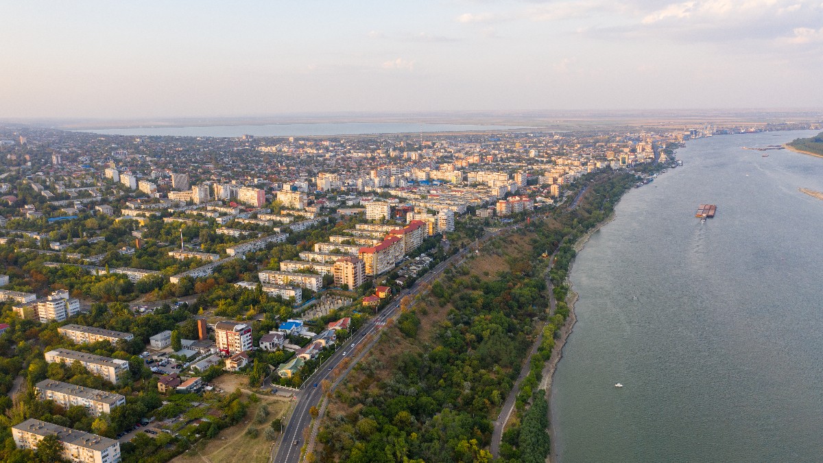 Imaginea surprinde o vedere aeriană spectaculoasă a orașului Galați, întins de-a lungul malului Dunării, cu numeroase blocuri, clădiri și spații verzi. În partea dreaptă se observă fluviul, pe care navighează o barjă, iar faleza și drumul de pe mal conturează peisajul urban.