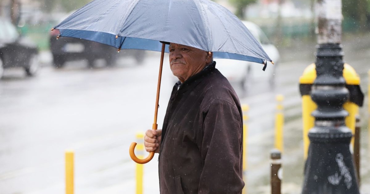 Un bărbat în vârstă, cu geacă maro și o umbrelă albastră pe care o ține deschisă, stă la semafor într-o zi ploioasă, marți, 27 mai 2025, București
