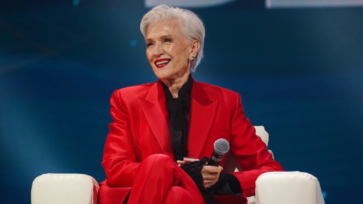 Maye Musk, mama lui Elon Musk, îmbrăcată într-un costum roșu, având un microfon în mână