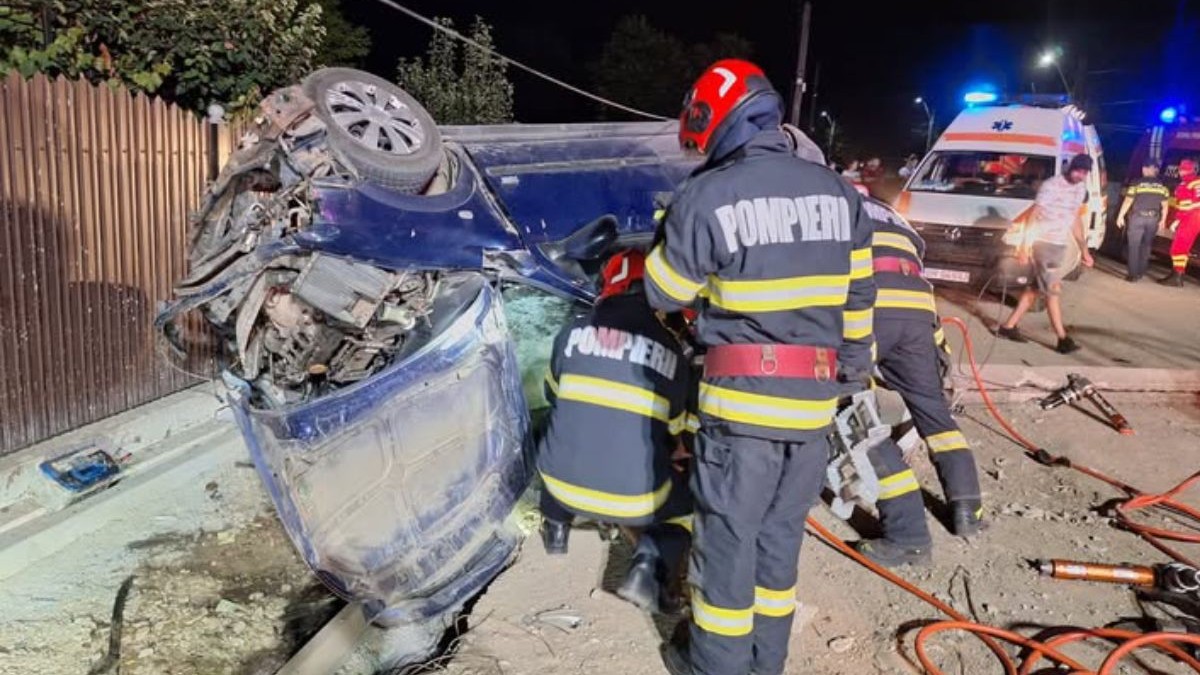 In fotografia de noapte se vad doi pompieri, unul in picioare, unul pe vine, langă o masină distrusa intr0un accident, in spare este o ambulanță, apamedici si oameni