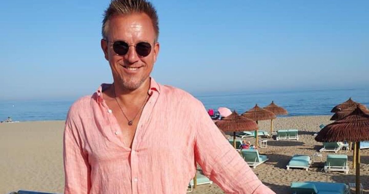 In imagine Anders Kurtén, CEO-ul Fraser Yachts,, intr-o cămașă lejeră roz pudrat, cu ochelari de soare, in spatele lui o plaja cu umbrele de stuf , nisip și marea