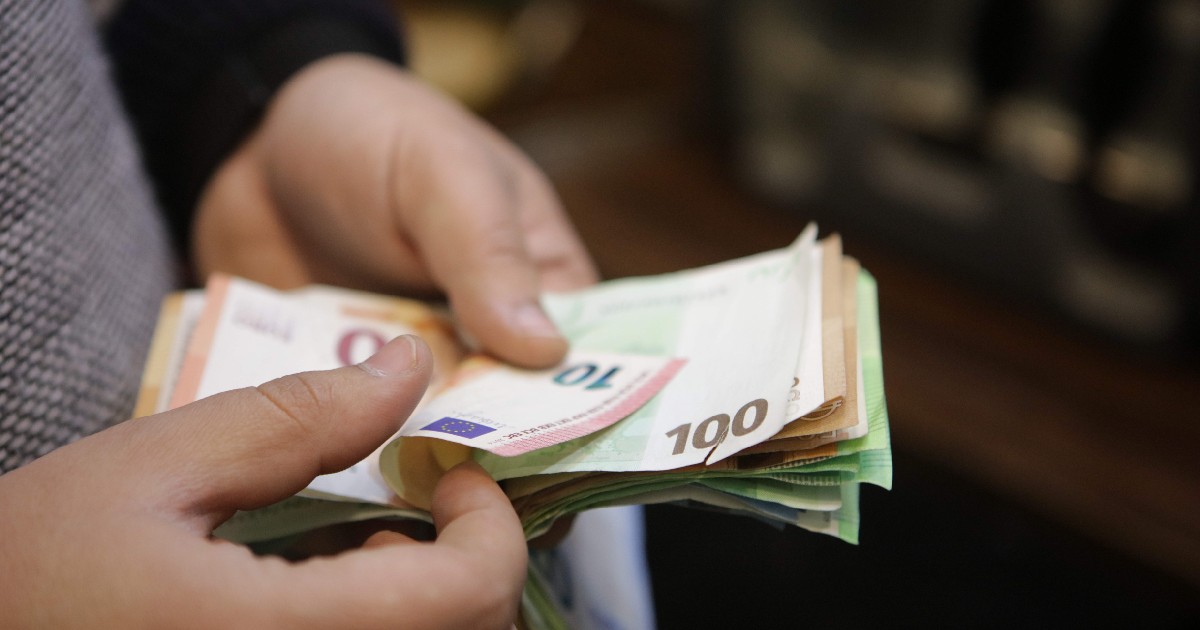 Imaginea surprinde planul apropiat al mâinilor unei persoane care răsfoiește un teanc de bancnote euro, fiind vizibile hartii de 10 și 100 de euro.