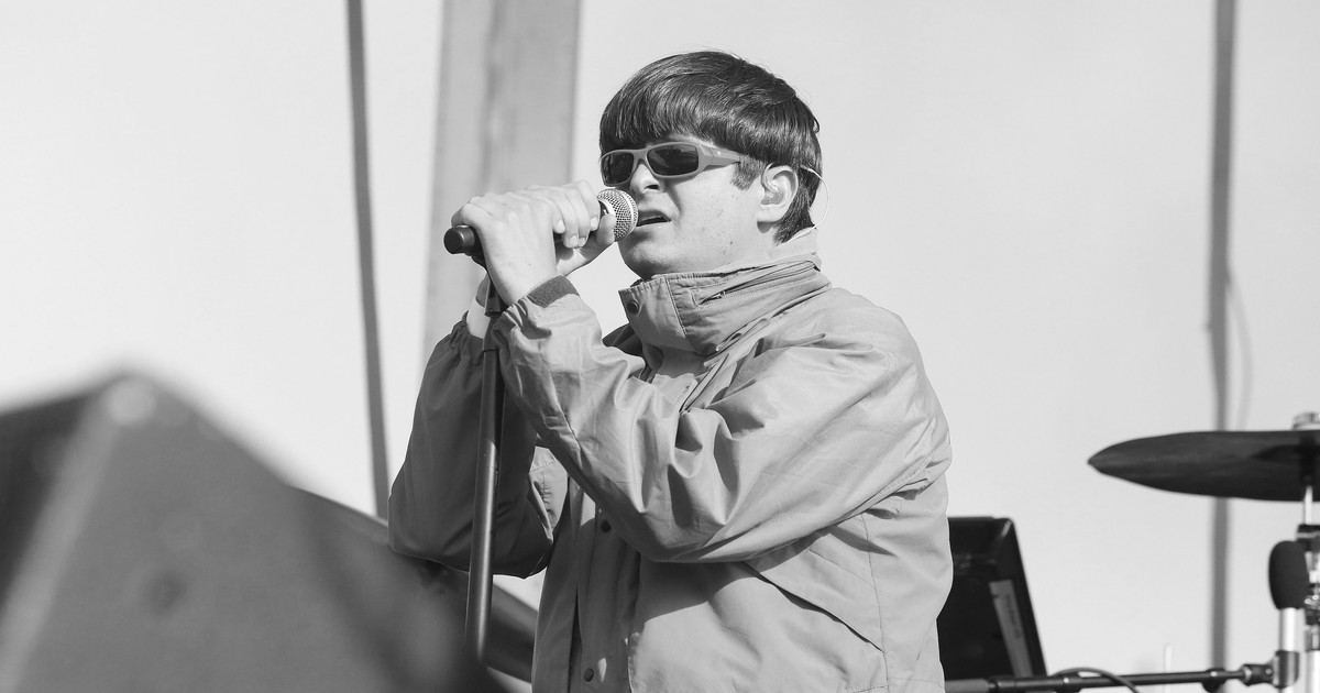Imaginea îl surprinde pe un artist în timpul unui concert, cântând la microfon pe o scenă în aer liber. Acesta poartă o geacă largă și ochelari de soare, având o apariție distinctivă și o atitudine concentrată în timpul prestației.