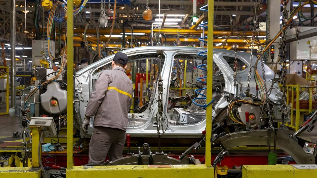 Fabrică mașini, unde în prim-plan, se vede un muncitor îmbrăcat în uniformă de protecție, lucrând la o caroserie metalică de autoturism aflată pe linia de producție.